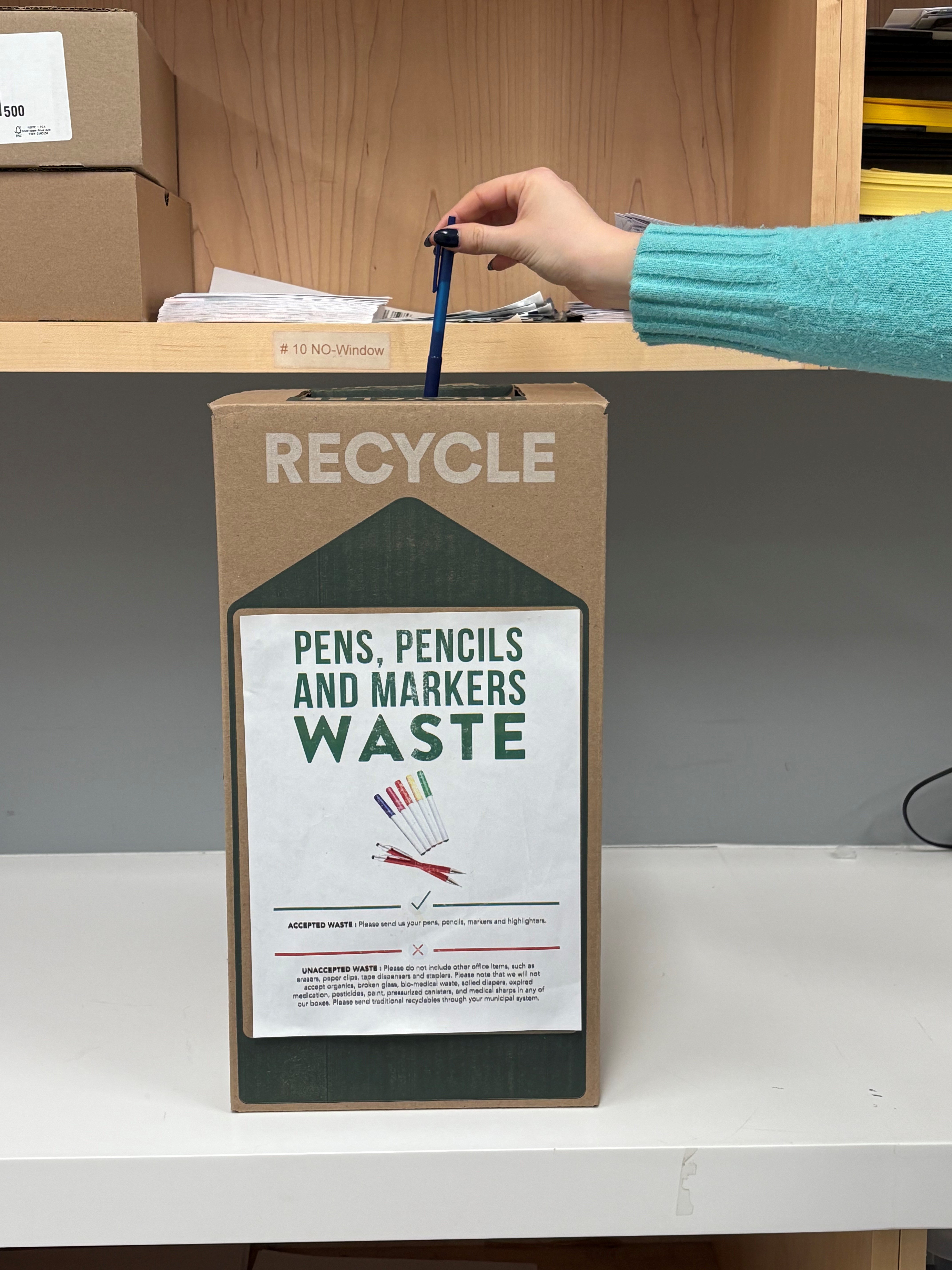 Pen being dropped into brown TerraCycle recycling box for pens, pencils and markers